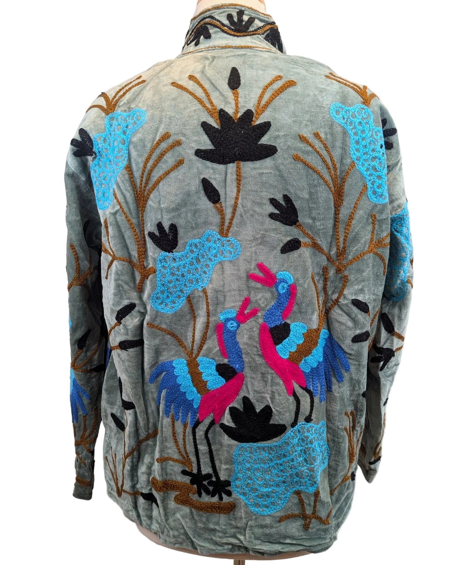 A vintage-style embroidered jacket displayed on a mannequin, showing the back view. The jacket has a muted sage green base with intricate embroidery of blue lotus flowers, brown branches, and two pink and blue birds.