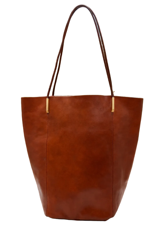 A large brown leather tote bag with double shoulder straps and gold-tone hardware, displayed on a round stone table with a blurred garden patio background.